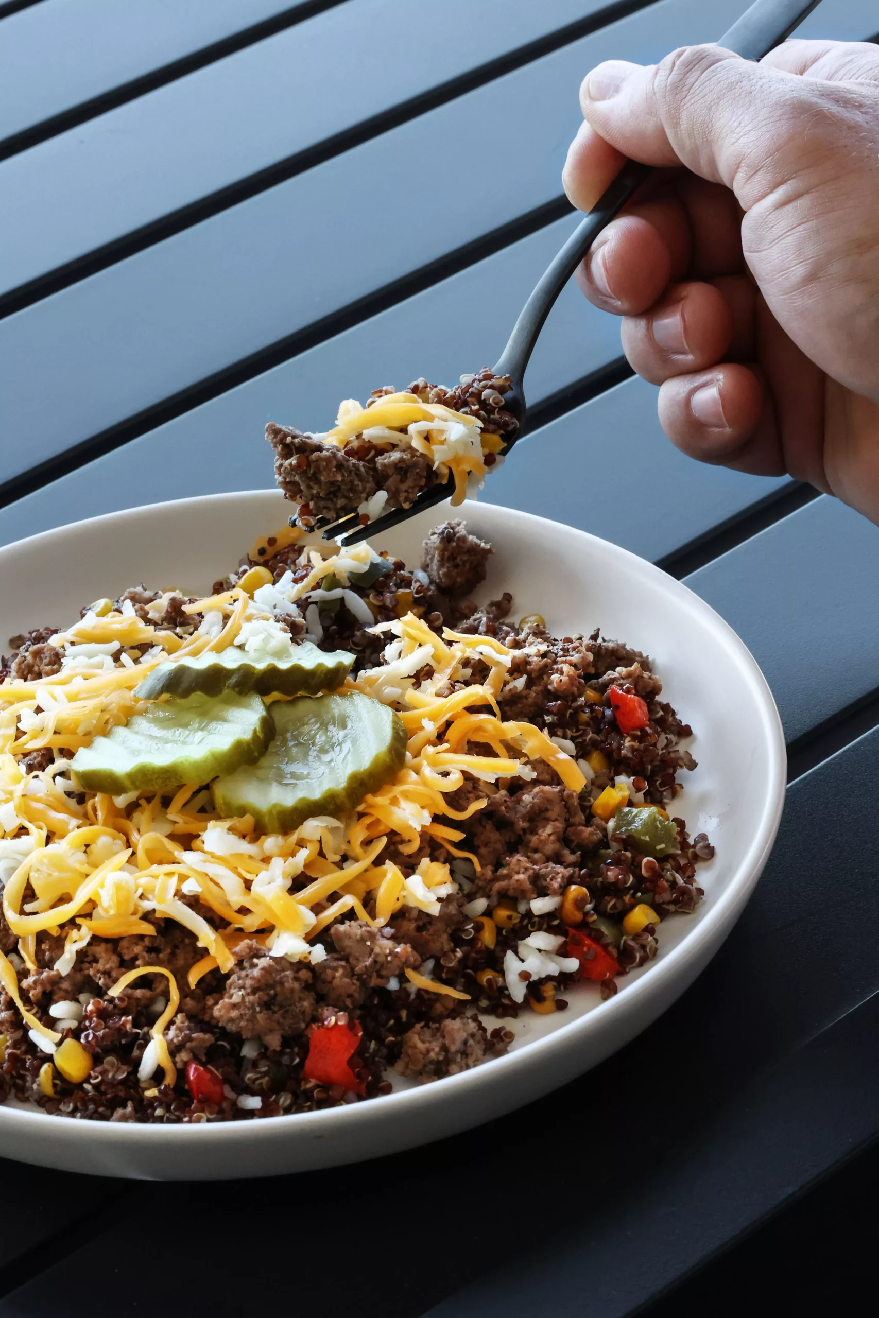 Person holding a fork over a cheeseburger-style bowl with ground beef, shredded cheese and pickles